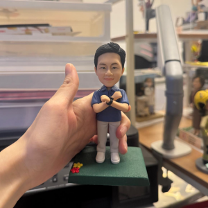 手持展示的公仔:角色穿藍色 POLO 衫、淺灰長褲與白色鞋,雙臂於胸前交叉,兩手腕配黑色手環/錶;腳下為綠色草地質感的矩形底座,前角點綴紅黃小花。背景為室內桌面與收納層架。