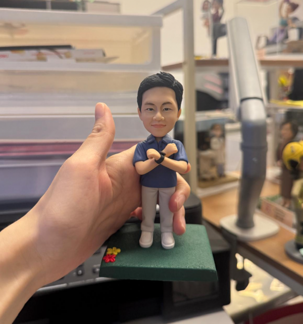 手持展示的公仔：角色穿藍色 POLO 衫、淺灰長褲與白色鞋，雙臂於胸前交叉，兩手腕配黑色手環／錶；腳下為綠色草地質感的矩形底座，前角點綴紅黃小花。背景為室內桌面與收納層架。