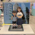 透明展示盒中的師生主題公仔：右側教師穿白色上衣與深藍長裙，手抱一本彩色封面的書；左側學生穿淺灰上衣與黑色短褲、白襪黑鞋，右手比 V 字手勢。腳下為灰色磚紋平台，兩側綠色草帶點綴小花；背板是藍天下的校舍外牆。
