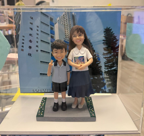 透明展示盒中的師生主題公仔：右側教師穿白色上衣與深藍長裙，手抱一本彩色封面的書；左側學生穿淺灰上衣與黑色短褲、白襪黑鞋，右手比 V 字手勢。腳下為灰色磚紋平台，兩側綠色草帶點綴小花；背板是藍天下的校舍外牆。