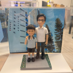 透明展示盒中的師生公仔：右側老師戴黑框眼鏡、穿白色有黑色滾邊的 POLO 衫與深色長褲；左側學生穿淺灰上衣與黑色短褲、白襪黑鞋，右手比 V 字手勢。腳下為灰色磚紋長方形平台，邊緣綠色草帶點綴小花；背板是藍天下的校舍立面與直排中文校名。