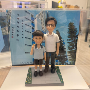 透明展示盒中的師生公仔：右側老師戴黑框眼鏡、穿白色有黑色滾邊的 POLO 衫與深色長褲；左側學生穿淺灰上衣與黑色短褲、白襪黑鞋，右手比 V 字手勢。腳下為灰色磚紋長方形平台，邊緣綠色草帶點綴小花；背板是藍天下的校舍立面與直排中文校名。