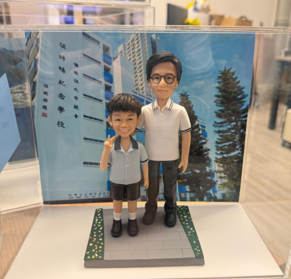 透明展示盒中的師生公仔：右側老師戴黑框眼鏡、穿白色有黑色滾邊的 POLO 衫與深色長褲；左側學生穿淺灰上衣與黑色短褲、白襪黑鞋，右手比 V 字手勢。腳下為灰色磚紋長方形平台，邊緣綠色草帶點綴小花；背板是藍天下的校舍立面與直排中文校名。