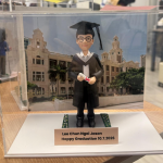 透明展示盒中的畢業公仔：角色戴黑色學士帽、穿黑色學袍配白色披肩，雙手捧著綁紅帶的卷軸；腳下為灰色磚紋平台，兩側綠色草帶點綴小花。背板為古典鐘樓與拱窗的校園建築。前方銅色銘牌黑字：「Lee Chun Ngai Jason／Happy Graduation 10.7.2026」。