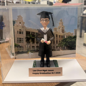 透明展示盒中的畢業公仔：角色戴黑色學士帽、穿黑色學袍配白色披肩，雙手捧著綁紅帶的卷軸；腳下為灰色磚紋平台，兩側綠色草帶點綴小花。背板為古典鐘樓與拱窗的校園建築。前方銅色銘牌黑字：「Lee Chun Ngai Jason／Happy Graduation 10.7.2026」。