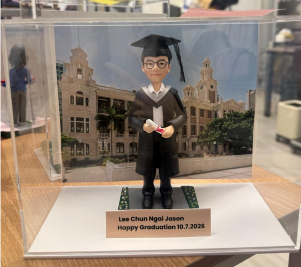透明展示盒中的畢業公仔：角色戴黑色學士帽、穿黑色學袍配白色披肩，雙手捧著綁紅帶的卷軸；腳下為灰色磚紋平台，兩側綠色草帶點綴小花。背板為古典鐘樓與拱窗的校園建築。前方銅色銘牌黑字：「Lee Chun Ngai Jason／Happy Graduation 10.7.2026」。
