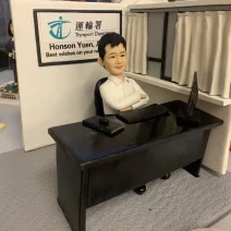 一件為運輸署上司 Honson Yuen 訂製的人像公仔榮休禮物。公仔身穿白恤衫，雙臂環抱，自信地坐在辦公桌前。背景牆上有運輸署的標誌和祝福語。