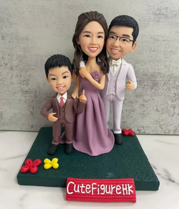 三口之家家庭人像公仔：媽媽穿紫色長洋裝站中間微笑，爸爸著淺灰西裝與領結站右側，孩子穿棕色小西裝站左側比出手勢；三人並肩站在深綠色草地質感底座上，周圍點綴紅色與黃色小花。前方紅色名牌寫有「CuteFigureHK」，背景為水泥紋灰牆與白色檯面。整體高雅溫馨，適合作為家庭人像公仔紀念禮物。