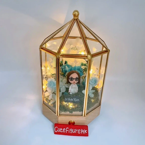 為女醫生 Dr. Vada Yuen 客製的人像公仔，公仔被放置在一個帶有燈串的黃銅玻璃罩中，周圍環繞著藍色和白色的保鮮花，是一款獻給醫護人員的、充滿治癒感的榮休禮物。