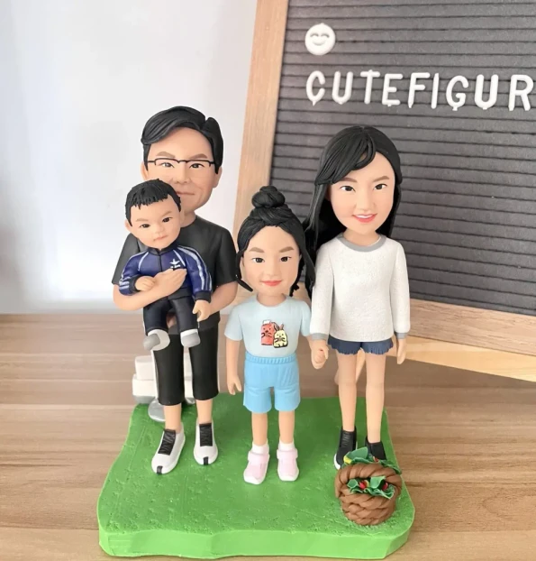 家庭人像公仔四人組：爸爸抱著穿深藍運動套裝的小朋友男孩站在左側；媽媽穿白色上衣與牛仔短褲、綠色鞋在右側；中間是紮高丸子頭、穿淺藍上衣與短褲的小女孩。四人站在草綠色不規則底座上，右前方放著棕色小籃子。背景為木質桌面與立式字母佈告板，上有「CUTeFIGUR…」字樣。整體清新溫馨，展現一家人的日常氛圍，適合作為小朋友主題的家庭人像公仔禮物與居家擺設。