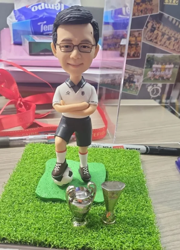 一位男士的足球主題榮休禮物人像公仔。他身穿球衣，腳踏足球，身旁有兩座獎盃。公仔站在仿真草皮底座上，背景有球隊照片，是一份紀念其貢獻的禮物。