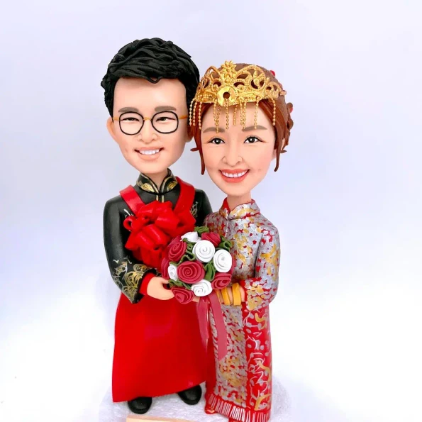 一張展示中式婚禮主題結婚公仔禮物的照片，圖中是一款為新人度身訂造的結婚公仔。新娘戴皇冠（鳳冠），身穿華麗裙褂；新郎則穿著傳統馬褂，並捧著花球。這款精緻的結婚公仔訂造作品，是充滿心思的新人結婚禮物、送好友結婚禮物或結婚一週年禮物的理想選擇。"