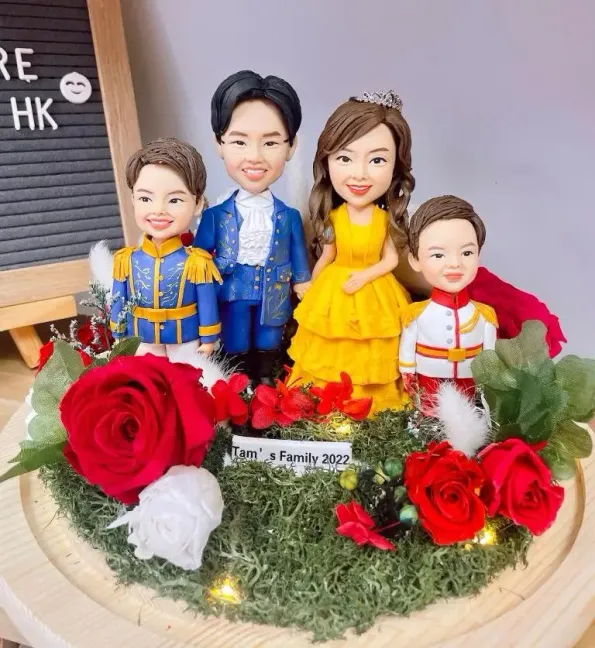四口之家主題家庭人像公仔擺設：爸爸穿藍色王子禮服、媽媽穿亮黃色層次長裙並配髮飾，兩名孩子分別穿藍金王子服與白紅小王子服。公仔站在苔蘚草地造景上，周圍點綴紅白玫瑰與小燈串，前方有「Family 2022」銘牌。背景為木紋底座與字母板，整體夢幻童話風格，適合作為家庭人像公仔禮物與居家展示。