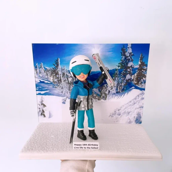 一位年輕人的18歲生日禮物運動公仔。他穿著全套滑雪裝備，在雪山背景前高舉雪板。名牌上寫著「Happy 18th Birthday. Live life to the fullest」。