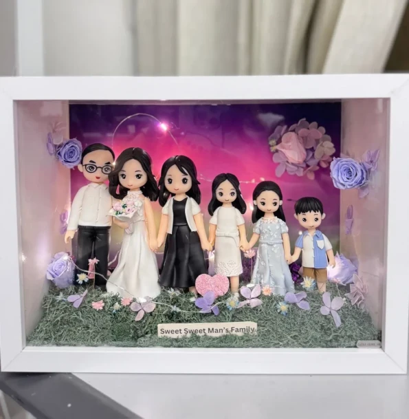 白色相框內的立體家庭場景：六位Q版人物公仔從左到右依序站立並牽手，包含戴眼鏡的爸爸、穿白色婚紗捧花的媽媽、兩位身穿黑白禮服的女孩、穿淡藍洋裝的女孩與穿藍白襯衫的小男孩。前景是草地與粉紫花朵，背景為紫粉漸層夜空搭配北極光效果與小燈串，角落點綴玫瑰花。