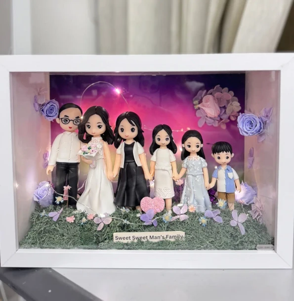 白色相框內的立體家庭場景：六位Q版人物公仔從左到右依序站立並牽手，包含戴眼鏡的爸爸、穿白色婚紗捧花的媽媽、兩位身穿黑白禮服的女孩、穿淡藍洋裝的女孩與穿藍白襯衫的小男孩。前景是草地與粉紫花朵，背景為紫粉漸層夜空搭配北極光效果與小燈串，角落點綴玫瑰花。