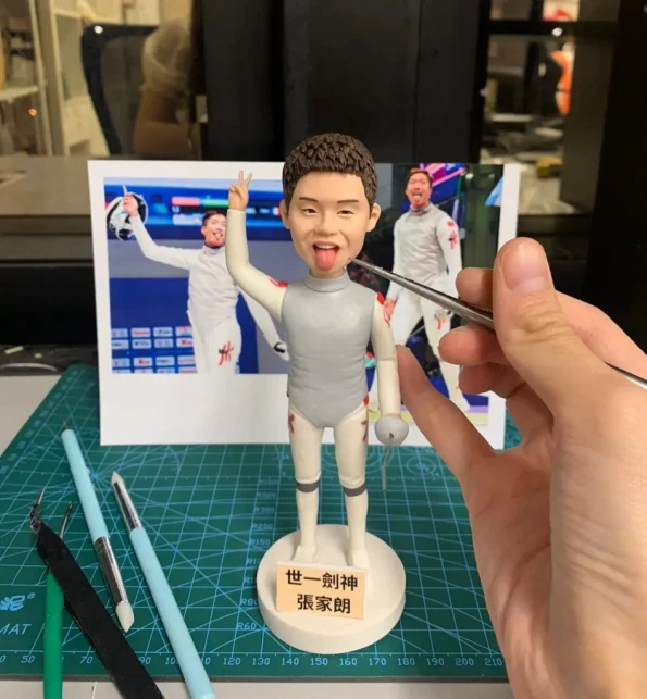 一個正在製作中的香港劍擊運動員張家朗人像公仔。公仔做出伸脷的V字手勢，底座寫著「世一劍神 張家朗」，背景是他的參考照片，展現了公仔的製作過程。