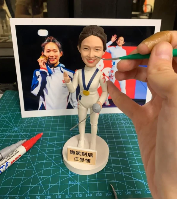 一個正在製作中的香港劍擊運動員江旻憓人像公仔。公仔掛著金牌，面帶燦爛微笑，底座寫著「微笑劍后 江旻憓」，背景是她的參考照片。