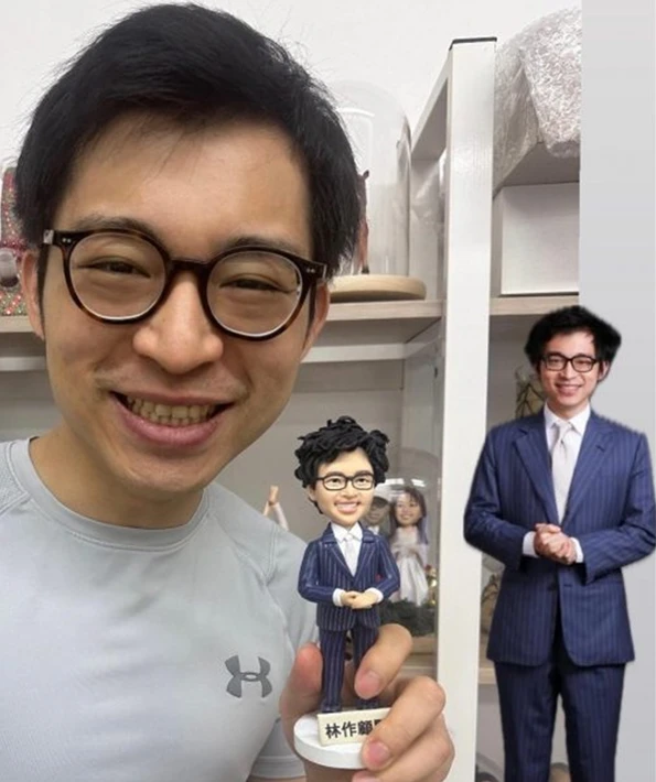 一張顧客開心展示他專屬訂製人像公仔的照片。圖中，一位戴眼鏡的男士手持一個穿著條紋西裝的迷你人像公仔，公仔底座上刻有名字；背景裡還有一張該西裝造型的真人照片作為對比，突顯了這款人像公仔的精緻還原度。