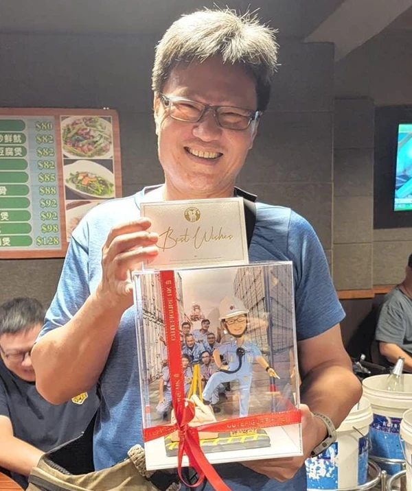 一位男士在榮休慶祝會上，欣慰地笑著，展示他收到的榮休禮物。這份禮物是一個客製化人像公仔，公仔以他穿著工作服的形象製作，背景更巧妙地融合了與團隊同事的合照，是一份極具紀念意義的禮物。