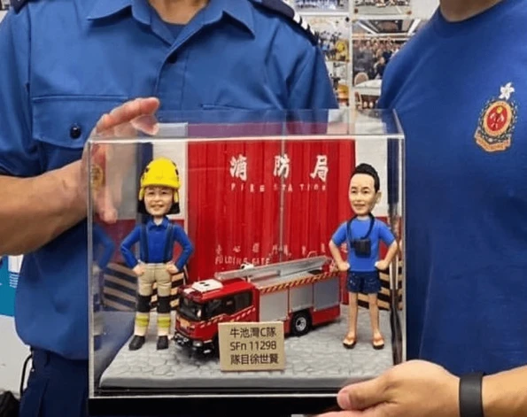 在消防局內，兩位消防員正捧著一份客製化的榮休或歡送禮物。這份禮物是一個精緻的立體場景，裡面有兩個消防員人像公仔、一輛消防車模型，背景是消防局的閘門，完美地呈現了這份消防公仔禮物的獨特性與紀念價值。