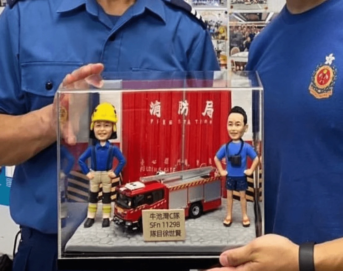 在消防局內，兩位消防員正捧著一份客製化的榮休或歡送禮物。這份禮物是一個精緻的立體場景，裡面有兩個消防員人像公仔、一輛消防車模型，背景是消防局的閘門，完美地呈現了這份消防公仔禮物的獨特性與紀念價值。
