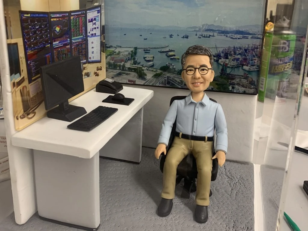 一個極具細節的客製化人像公仔，呈現一位戴眼鏡的男士面帶微笑坐在他的辦公室場景中。這個場景完美還原了他的專業工作環境，包括一張擺放著電腦和電話的辦公桌，背景是多個顯示著金融數據的螢幕，以及一扇能看到海港景色的窗戶。這是一份極具心思的榮休禮物。