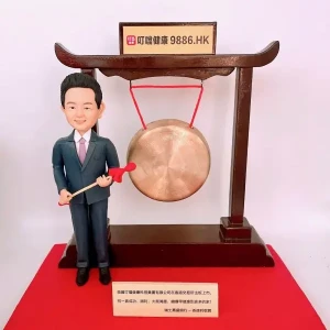 一隻男士人像公仔身穿西裝，手持紅色鼓槌，站在大鑼旁邊。背景設有公司上市敲鑼裝置，上方牌匾寫有公司名稱及股票代碼「9886.HK」。公仔和裝置放置於紅色底座上。這是特別為公司上市設計的人像公仔禮物。