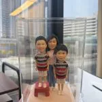 透明展示盒內的三口之家家庭人像公仔：媽媽站在後方中央，兩位小男孩站在前方並肩；左側孩子踩著紅色小凳增高。三人皆穿著休閒條紋上衣與短褲、白色鞋子，置於米白色方形底座。背景為辦公室窗景與高樓外觀，光線明亮，整體呈現溫馨可愛的家庭人像公仔禮物擺設。