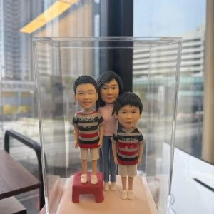 透明展示盒內的三口之家家庭人像公仔：媽媽站在後方中央，兩位小男孩站在前方並肩；左側孩子踩著紅色小凳增高。三人皆穿著休閒條紋上衣與短褲、白色鞋子，置於米白色方形底座。背景為辦公室窗景與高樓外觀，光線明亮，整體呈現溫馨可愛的家庭人像公仔禮物擺設。