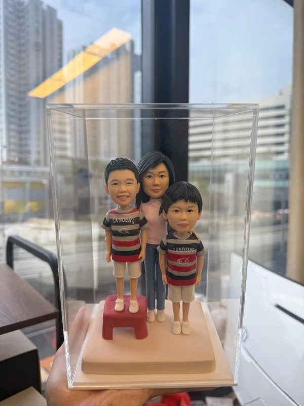 透明展示盒內的三口之家家庭人像公仔：媽媽站在後方中央，兩位小男孩站在前方並肩；左側孩子踩著紅色小凳增高。三人皆穿著休閒條紋上衣與短褲、白色鞋子，置於米白色方形底座。背景為辦公室窗景與高樓外觀，光線明亮，整體呈現溫馨可愛的家庭人像公仔禮物擺設。