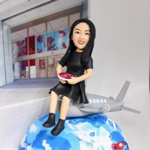 一件為即將退休移民的女士訂製的人像公仔禮物。公仔手持DFS標誌，坐在一架停在地球模型上的飛機上，象徵著她告別事業，開啟全新的海外生活。