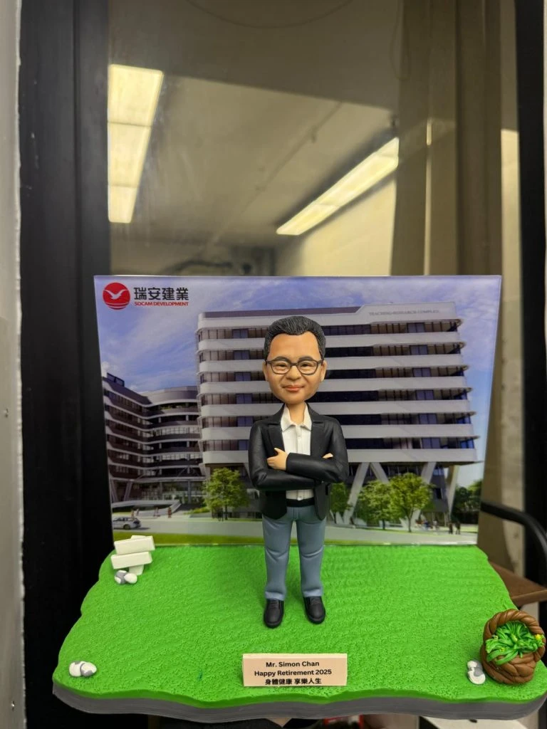 一個為瑞安建業(SOCAM)的陳先生(Mr. Simon Chan)訂製的2025年榮休禮物。模型主角是一位雙手抱胸的人像公仔，他自信地站在以公司建築項目為背景的展板前。公仔腳下的草地場景配有菜籃，寓意健康生活，是一份祝福其開啟人生新篇章的工程師禮物。