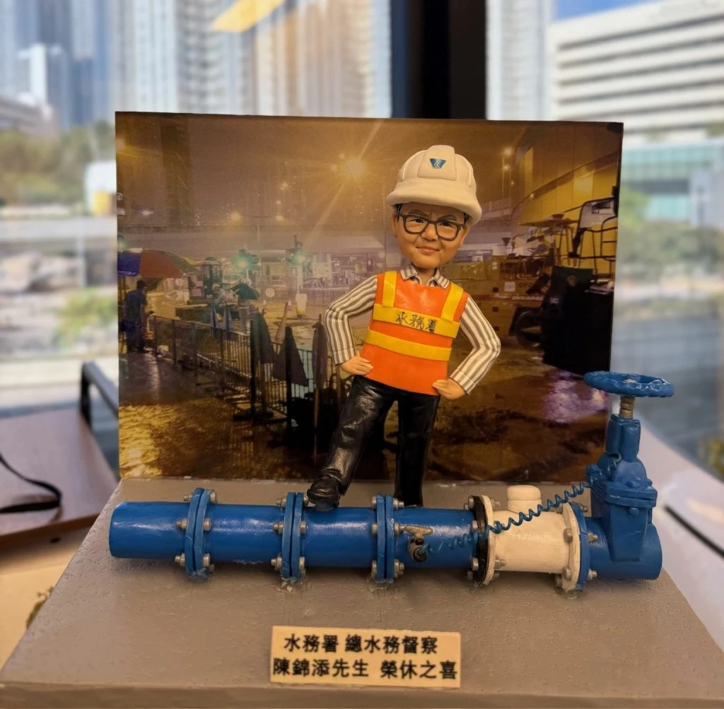 一個為香港水務署總水務督察陳錦添先生訂製的榮休禮物。模型主角是一個戴著安全帽和水務署反光衣的人像公仔，他充滿自信地單腳踏在精緻的藍色水管模型上。這份獨特的工程師禮物，背景是一張夜間工程照片，銘牌上刻有榮休祝福字句。