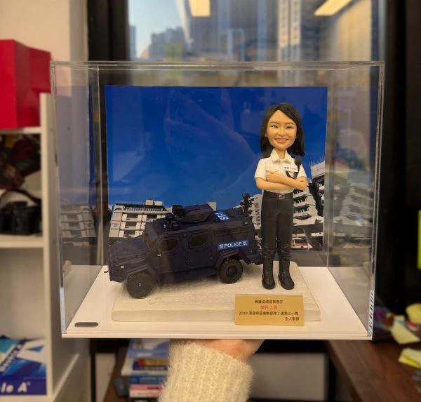 一個裝在透明展示盒中的警察榮升禮物。禮物主體是一個女性總督察的客製化人像公仔，她面帶微笑、雙手抱胸，身旁停著一輛精細的警隊裝甲車模型。前方的銘牌顯示這份禮物是由其PTU舊同袍贈送，祝賀她榮升之喜。