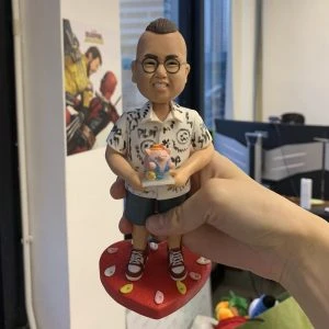 一個戴眼鏡、穿花紋襯衫和短褲的人像公仔,手中拿著小豬造型蛋糕,站在紅色愛心底座上,背景為辦公室環境。