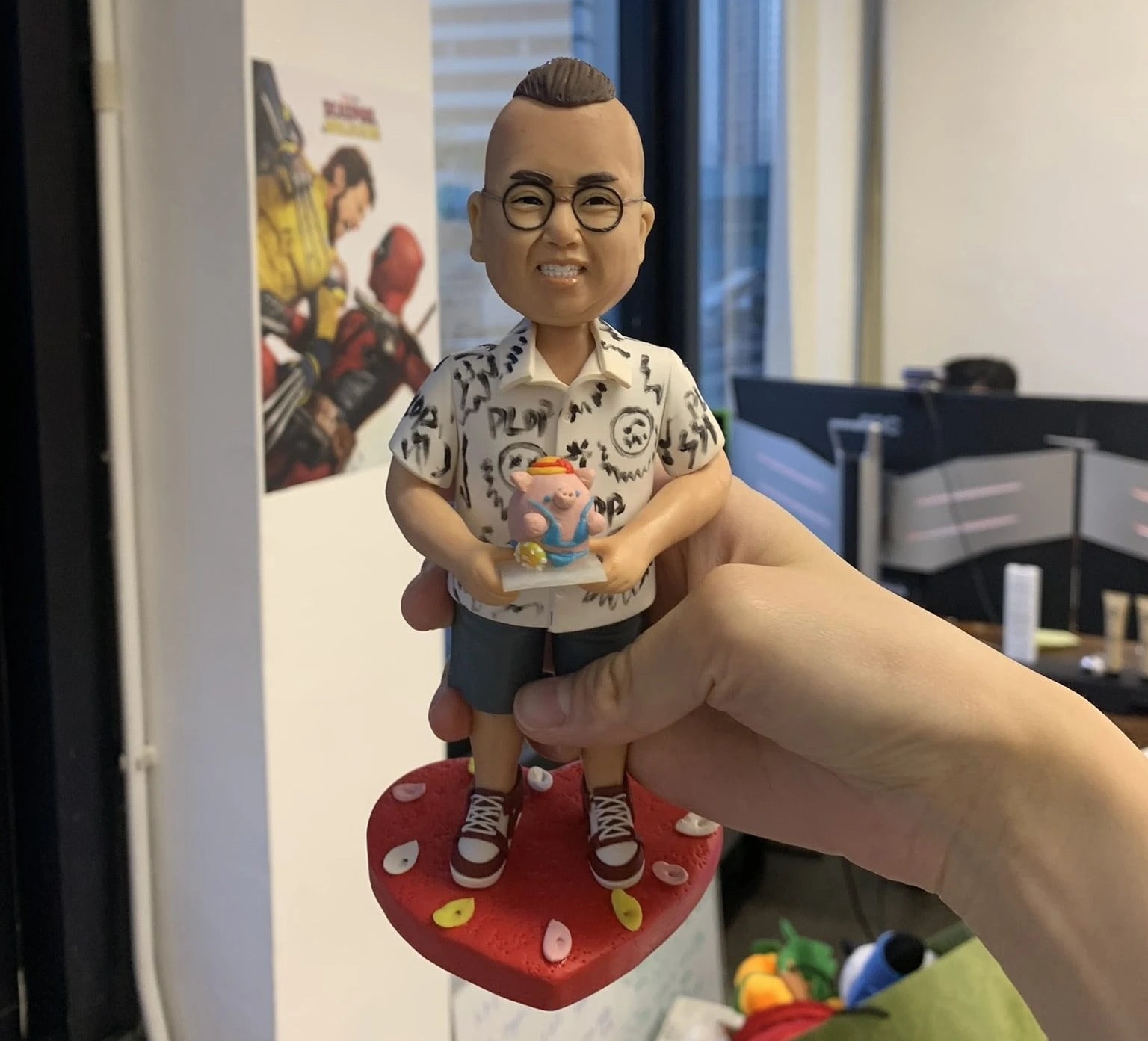 一個戴眼鏡、穿花紋襯衫和短褲的人像公仔，手中拿著小豬造型蛋糕，站在紅色愛心底座上，背景為辦公室環境。