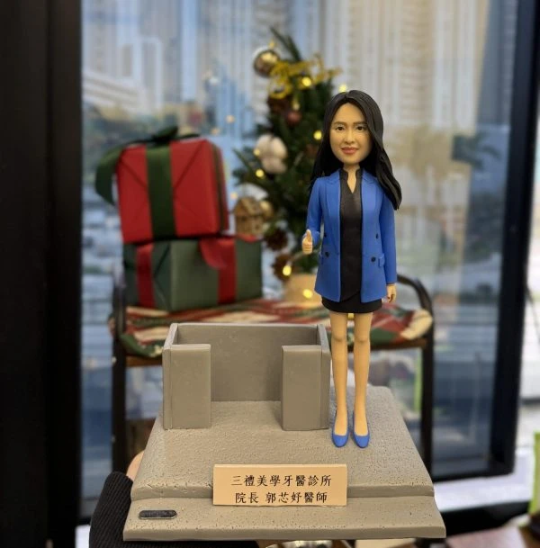 一張為「三禮美學牙醫診所」院長郭芯妤醫師訂製的人像公仔照片。圖中郭醫師的公仔身穿藍色外套，面帶微笑並豎起大拇指，站在刻有其名銜的專屬底座上。這款人像公仔是祝賀開張禮物的最佳選擇，也極適合作為上司榮升、公司週年慶的賀禮，甚至在公司上市等重要時刻，用以表彰核心人物。