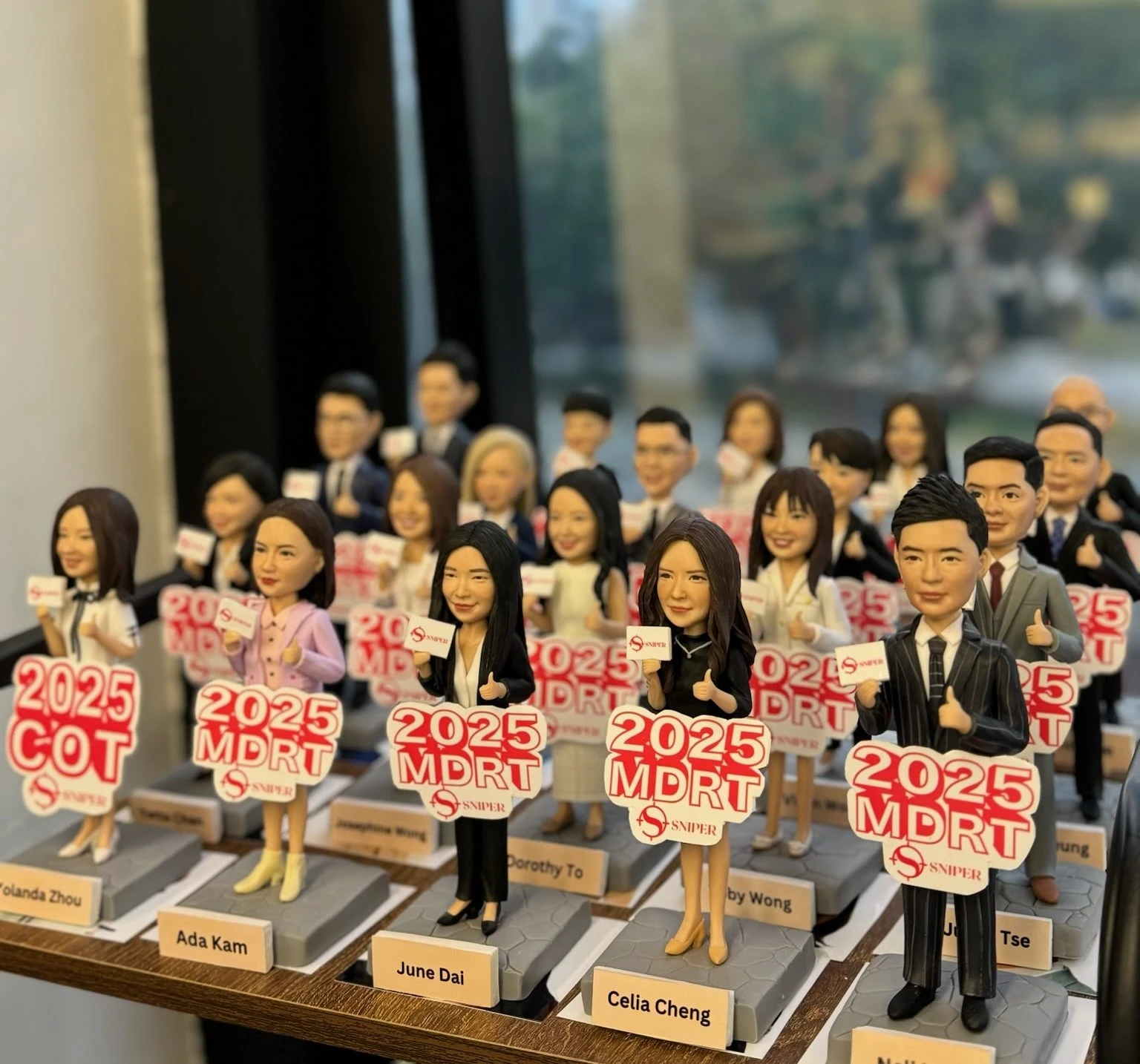 一張展示著一支精英團隊人像公仔的照片。圖中數十個根據團隊成員樣貌訂製的公仔整齊排列，每位成員都手持著『2025 MDRT』的目標牌。這款極具氣勢的團隊禮物，非常適合用於公司週年慶、激勵團隊，也可用於祝賀公司上市或上司榮升。