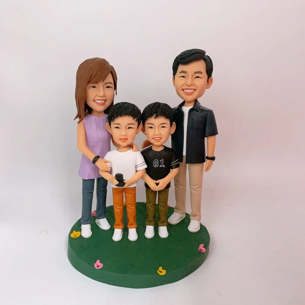 四口之家家庭人像公仔：爸爸穿深色外套與米色長褲站右側微笑，媽媽著淡紫色無袖上衣站左側；兩位孩子站在中間並肩而立，一位穿黑色上衣與卡其褲，另一位穿白色上衣與橘色長褲手握小麥克風。全員腳穿白鞋，置於圓形草綠底座，上面散落粉、黃小花裝飾；背景為純白牆面。整體活潑溫馨，適合作為家庭人像公仔禮物與居家擺設。