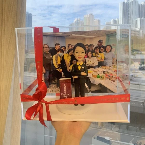 一份用紅色絲帶包裝的客製化榮升/調職禮物。禮物主體是一個女性人像公仔，她穿著制服、雙手抱胸，身旁有「九龍灣」的標示牌；背景是一張溫馨的同事派對大合照，整個設計充滿心思。