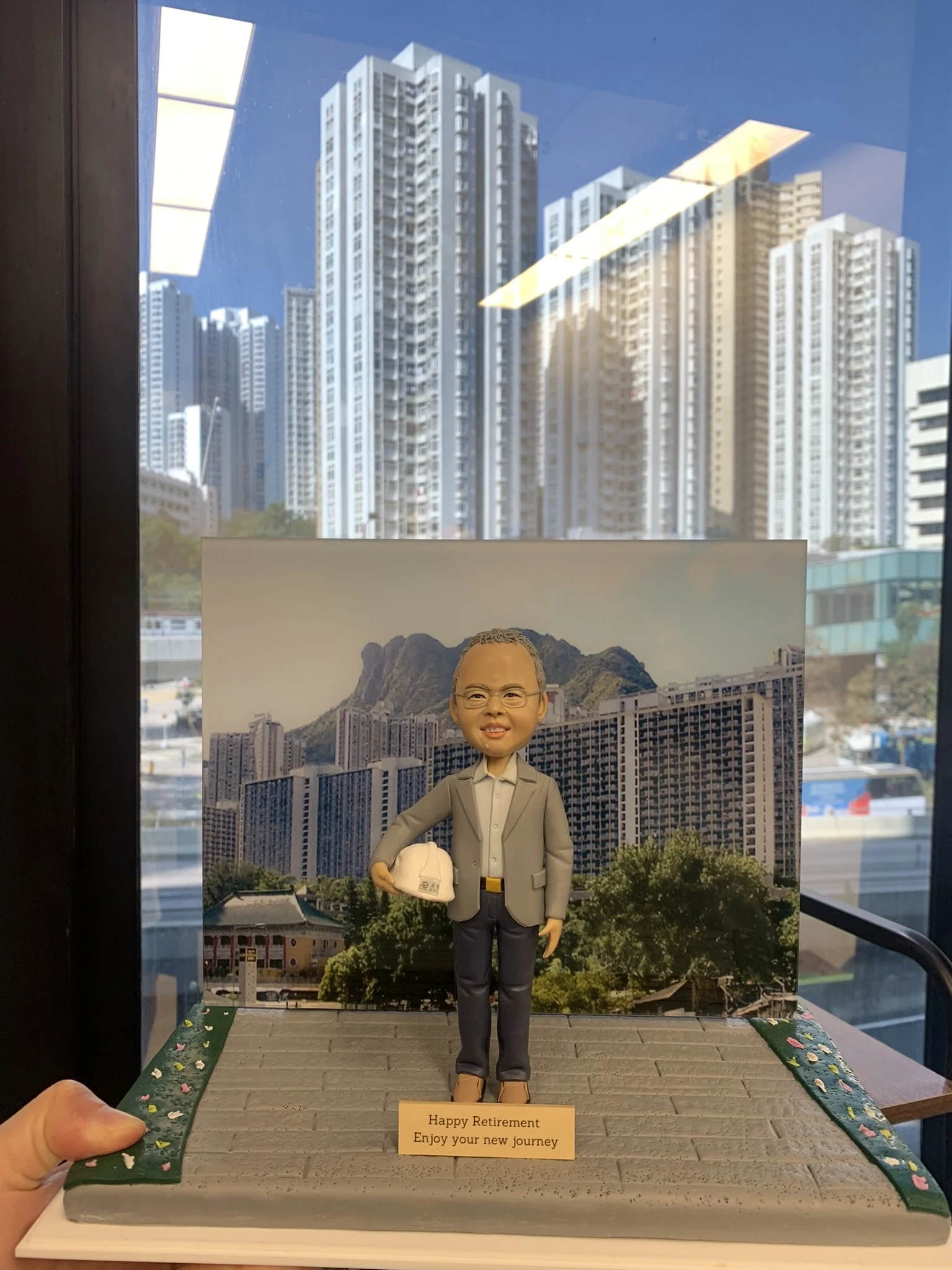 一個以香港獅子山為背景的榮休禮物。模型主角是一位戴眼鏡、身穿西裝、手持安全帽的人像公仔。他站在以獅子山和周邊住宅樓宇為背景的展板前，是一份向建設社區的工程師致敬的禮物，銘牌上寫有退休祝福。