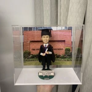 一位男士的碩士畢業人像公仔。他身穿畢業袍，站在香港大學的背景相片前，底座寫有「Master of Philosophy, Class of 2024」，是一份隆重的畢業禮物。