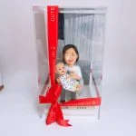 透明展示盒中的母子家庭人像公仔：短髮媽媽身穿白色上衣、淺色長褲，懷抱著穿白底黑點連身衣的嬰孩；底座前方有金屬感銘牌刻「2023」。展示盒外繫有紅色絲帶與蝴蝶結，側邊印字裝飾增添儀式感。背景為室內白牆與窗簾，整體溫馨可愛，適合作為家庭人像公仔禮物與收藏擺設。
