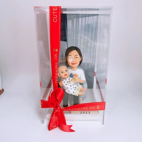 透明展示盒中的母子家庭人像公仔：短髮媽媽身穿白色上衣、淺色長褲，懷抱著穿白底黑點連身衣的嬰孩；底座前方有金屬感銘牌刻「2023」。展示盒外繫有紅色絲帶與蝴蝶結，側邊印字裝飾增添儀式感。背景為室內白牆與窗簾，整體溫馨可愛，適合作為家庭人像公仔禮物與收藏擺設。