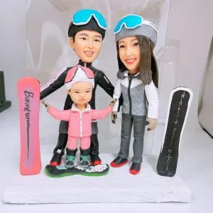 冬季滑雪主題的家庭人像公仔三人組:大人兩位分別穿黑色與灰白色滑雪服,頭戴護目鏡與帽子;中間是穿粉色連身雪服的小朋友,腳踩滑雪板站在綠白相間的雪地基座上。左右兩側各豎立一塊滑雪板模型(左為粉色字樣板、右為黑灰色板)。整體置於白色台座與簡潔背景前,呈現一家人在雪場合影的運動風格擺設,適合作為家庭人像公仔禮物。