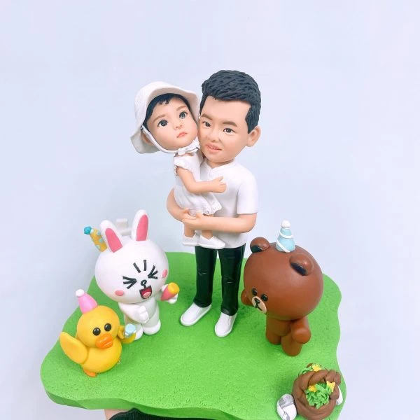 爸爸抱著戴白色遮陽帽的幼童站在草地造景上的家庭人像公仔；父子皆穿白色上衣、爸爸穿深綠褲與白鞋。周圍擺放派對氛圍的小動物公仔：黃色小鴨、戴派對帽的白兔與棕熊，以及野餐籃與小蛋糕裝飾。整體色彩清新童趣，適合作為家庭人像公仔禮物與生日派對主題擺設。
