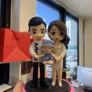 手掌捧著的小型家庭人像公仔：爸爸穿白襯衫與黑褲，媽媽著米白色洋裝，兩人合抱懷中的嬰兒公仔（戴藍色漁夫帽、穿藍白條紋衣）站在黑色圓台上，外圈以粉紅色邊框點綴。背景為室內辦公室窗景與紅色紙袋，整體溫馨可愛，適合作為家庭人像公仔紀念禮物。
