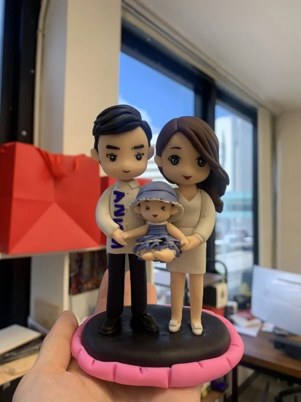 手掌捧著的小型家庭人像公仔：爸爸穿白襯衫與黑褲，媽媽著米白色洋裝，兩人合抱懷中的嬰兒公仔（戴藍色漁夫帽、穿藍白條紋衣）站在黑色圓台上，外圈以粉紅色邊框點綴。背景為室內辦公室窗景與紅色紙袋，整體溫馨可愛，適合作為家庭人像公仔紀念禮物。