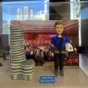 一款為興聯集團的資深工程師Nelson Wong訂製的退休禮物人像公仔，被封存在透明展示盒中。公仔身穿工程制服、手持安全帽，自信地站在一座獨特的建築模型旁，背景是公司團隊在「魯班先師寶誕」活動上的大合照，象徵著對其畢生貢獻的最高敬意。